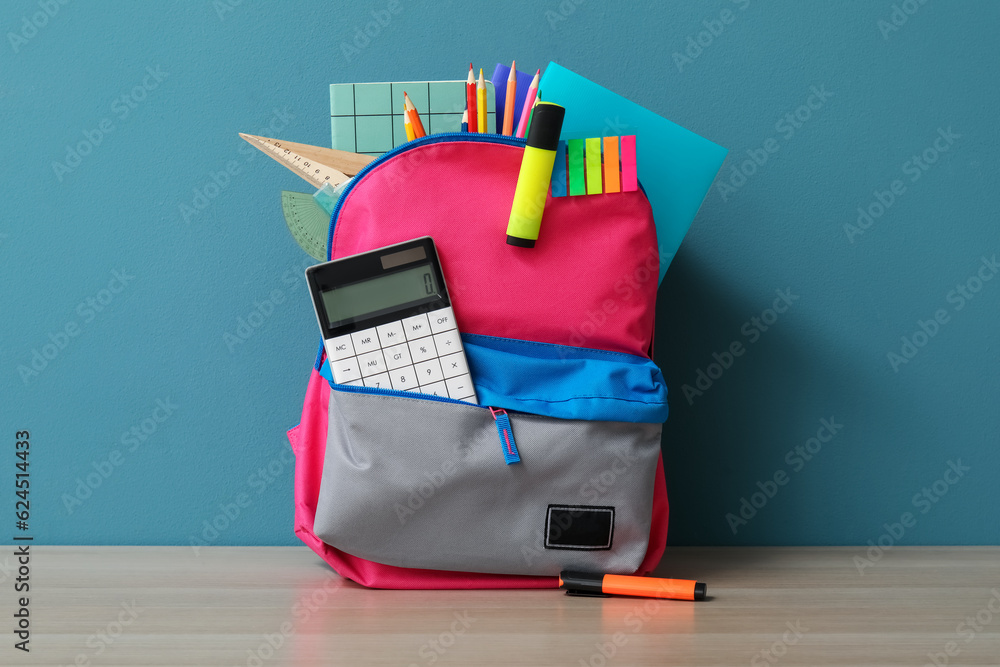 Colorful school backpack with different stationery and calculator on wooden table near blue wall