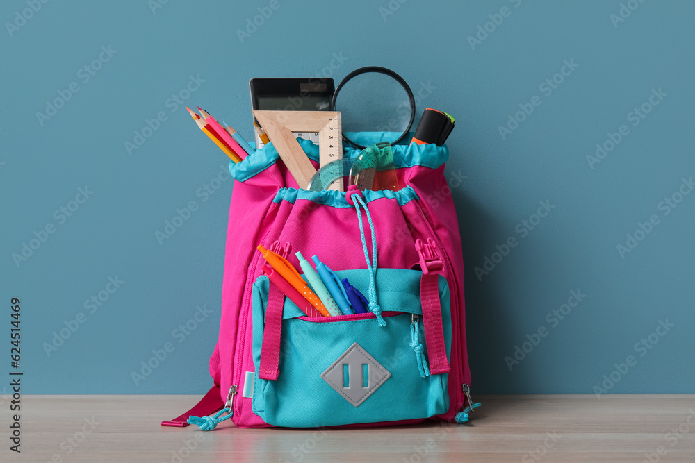 Colorful school backpack with different stationery and magnifier on wooden table near blue wall