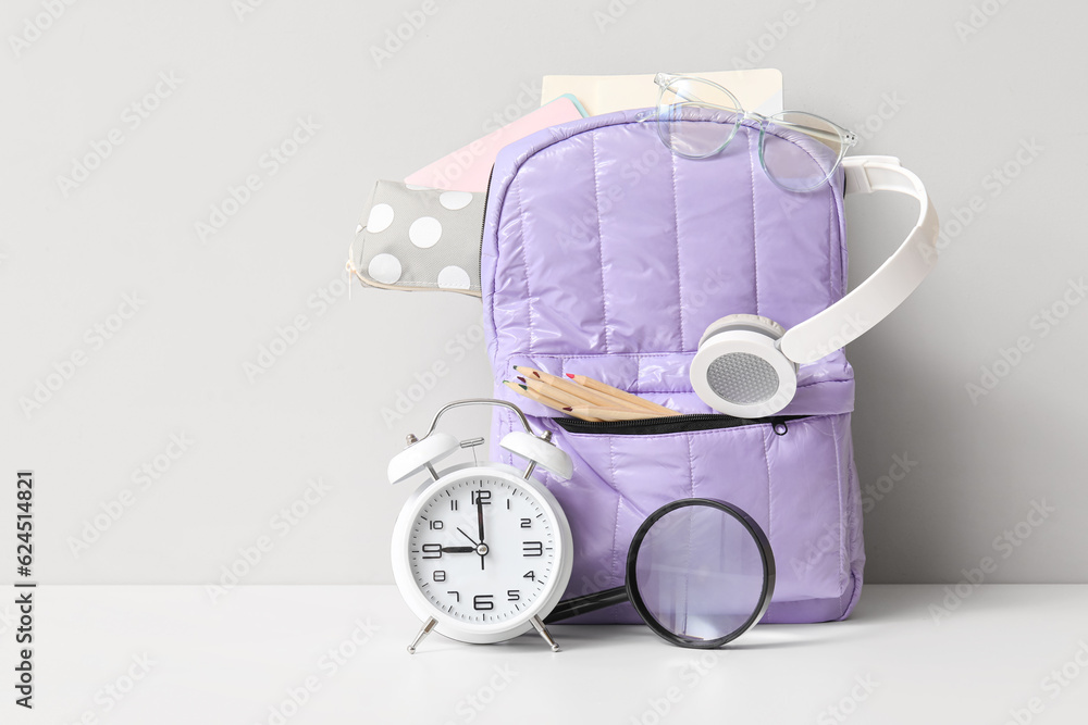 Purple school backpack with different stationery and alarm clock on white table near wall