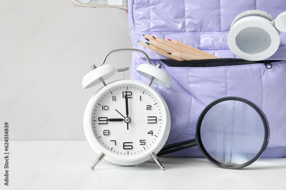 Purple school backpack with stationery and alarm clock on white table near wall