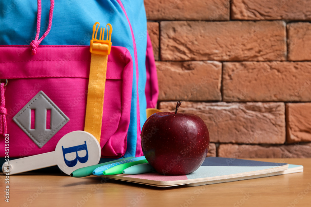 Colorful school backpack with different stationery and fresh apple on wooden table near brown brick 