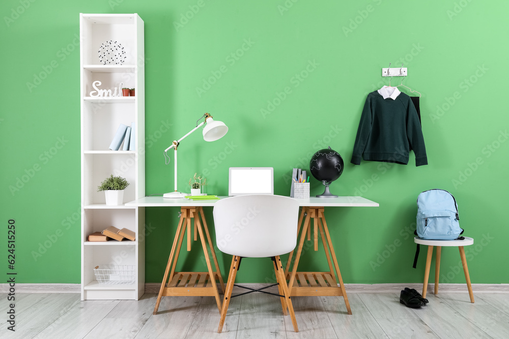 Workplace with desk, shelving unit, laptop computer, backpack and stylish school uniform hanging on 