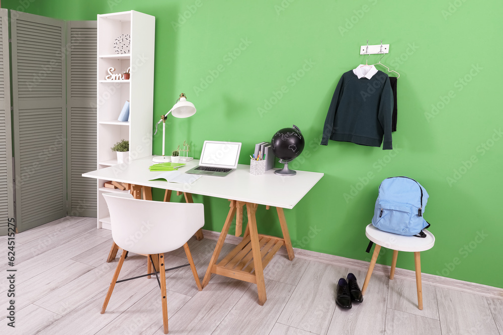 Workplace with desk, shelving unit, laptop computer, backpack and stylish school uniform hanging on 