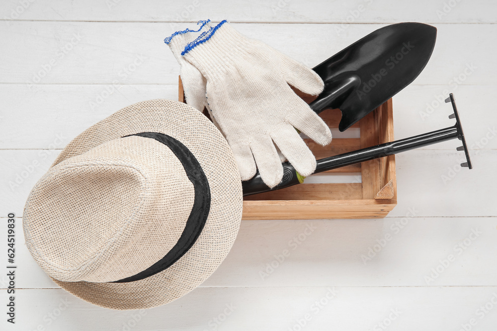 Box with different gardening tools and hat on light wooden background