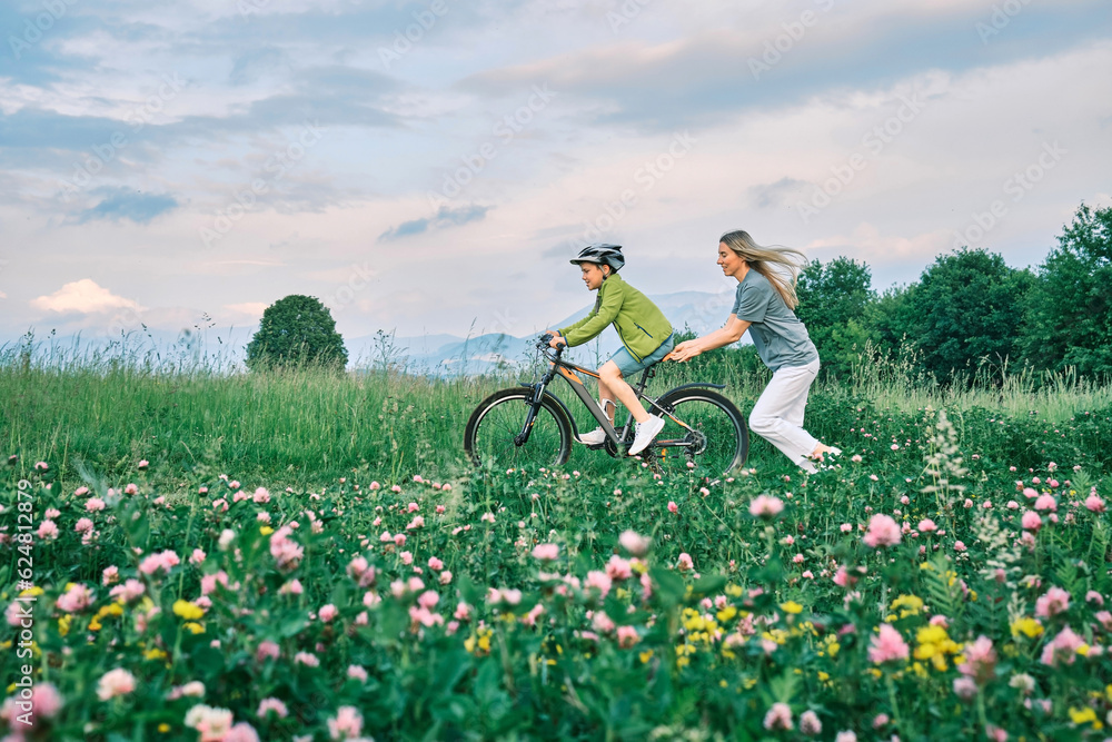 Mother teaching son to ride bicycle. Happy cute boy in helmet learn to riding a bike in park on gree