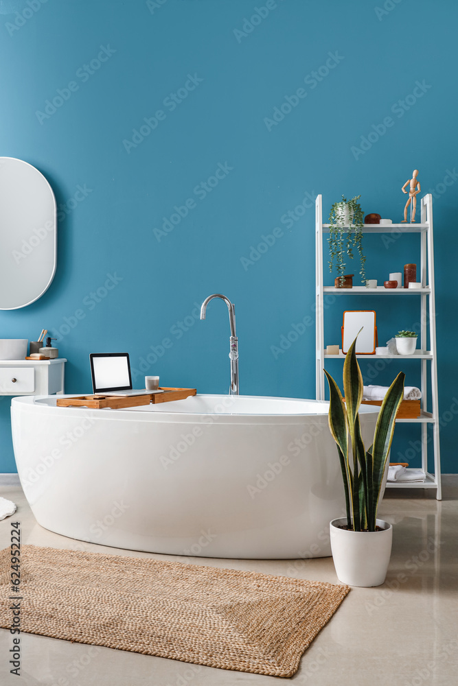 Interior of bathroom with bathtub, modern laptop and shelving unit