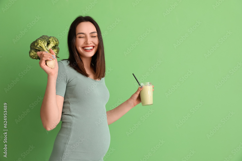 Young pregnant woman with glass of smoothie and broccoli on green background