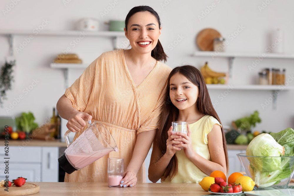 Young woman with her little daughter pouring fresh smoothie into glass in kitchen