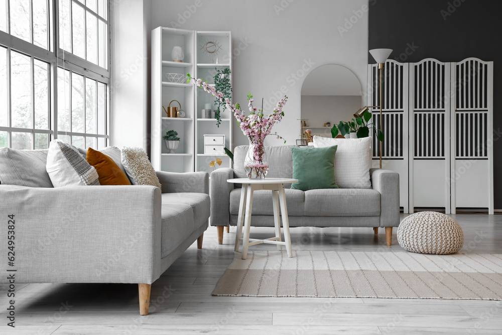 Interior of living room with grey sofas and blooming sakura branches on coffee table near big window