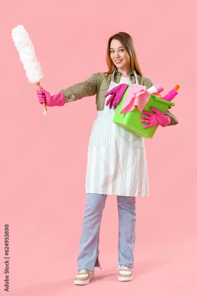 Young woman with pp-duster and cleaning supplies on pink background