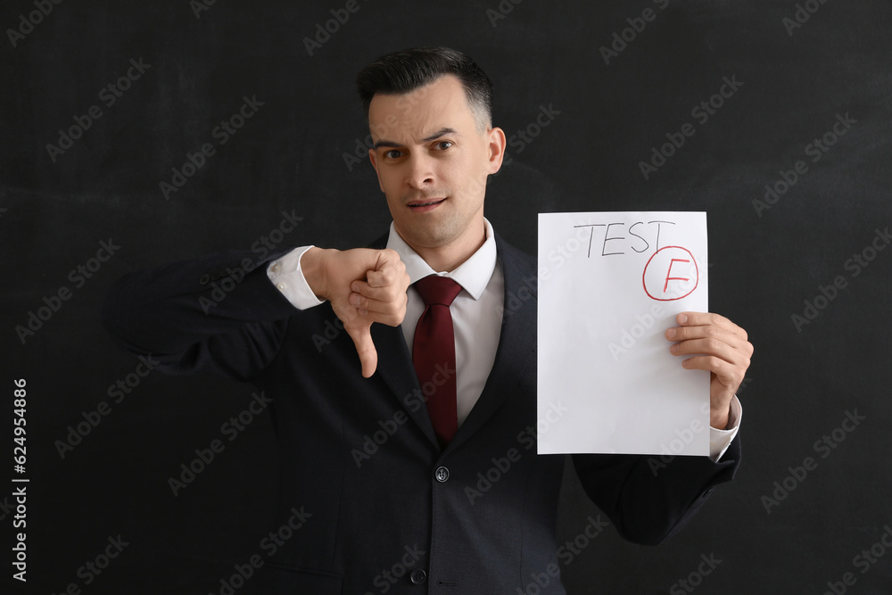 Male teacher with checked test sheet showing thumb-down near blackboard in classroom