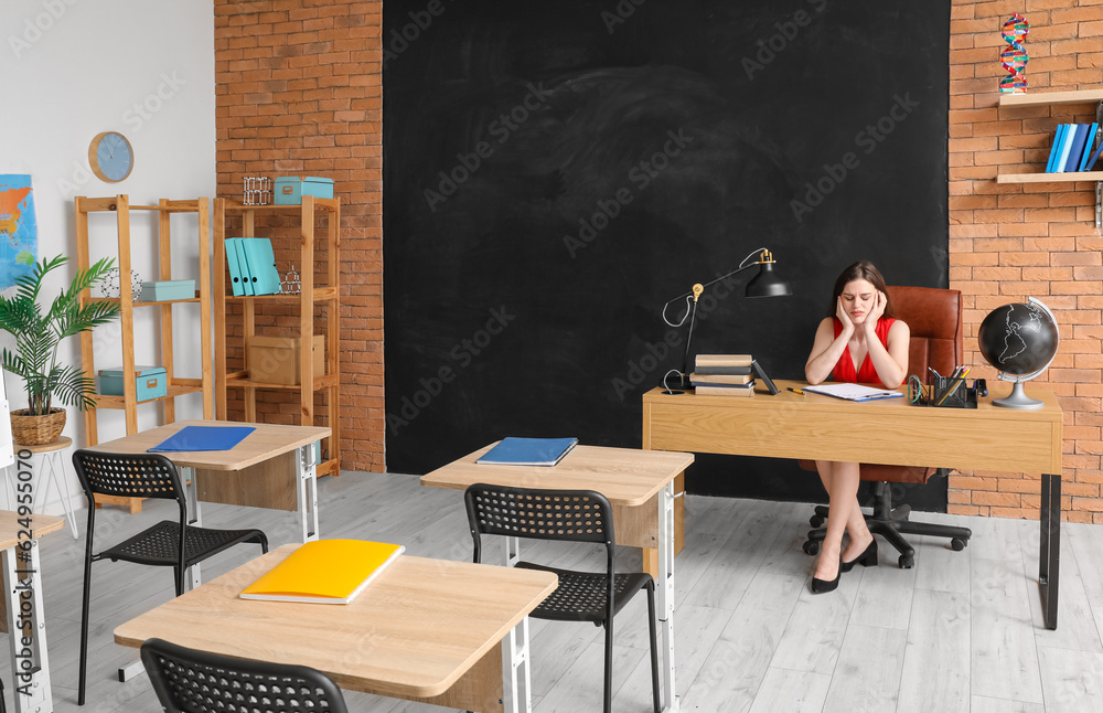 Tired female teacher sitting at table in classroom