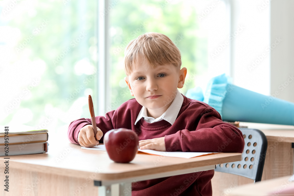 Happy little boy with blue school cone sitting at desk in classroom