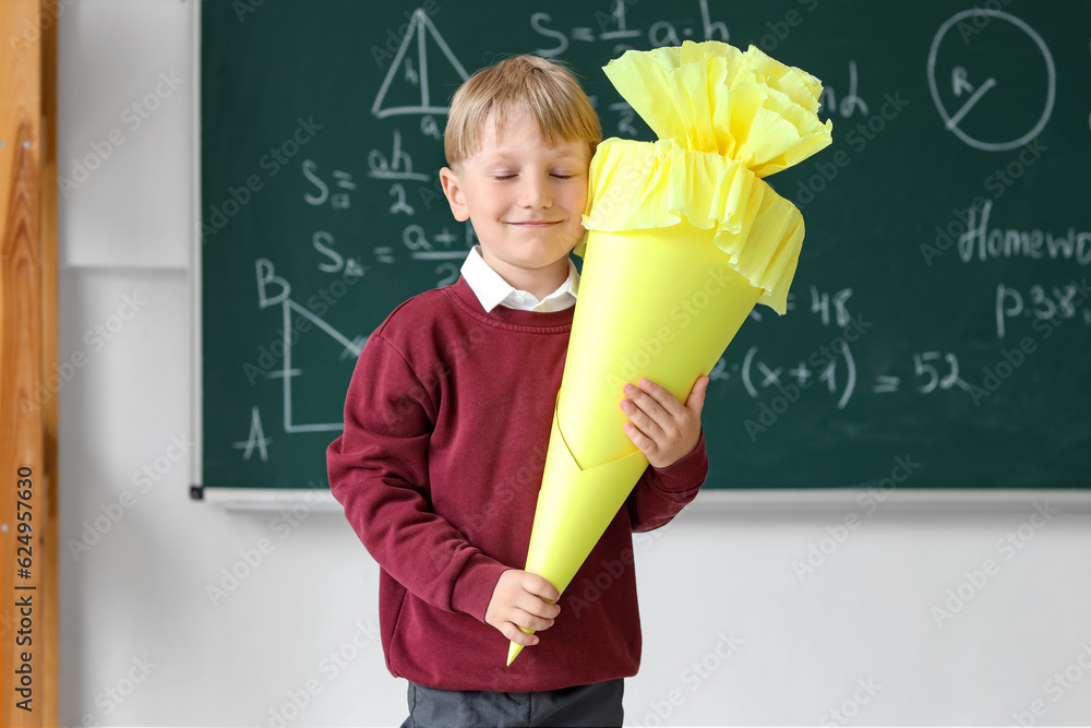 Happy little boy with yellow school cone in classroom near blackboard