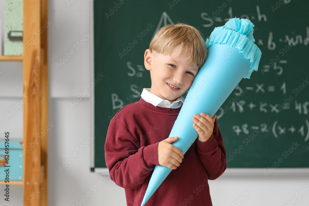 Happy little boy with blue school cone in classroom near blackboard