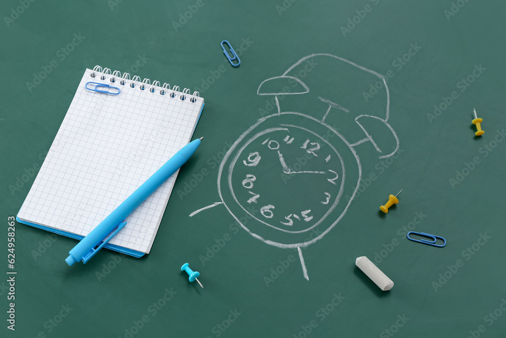 Blank notebook with stationery and drawn alarm clock on green chalkboard