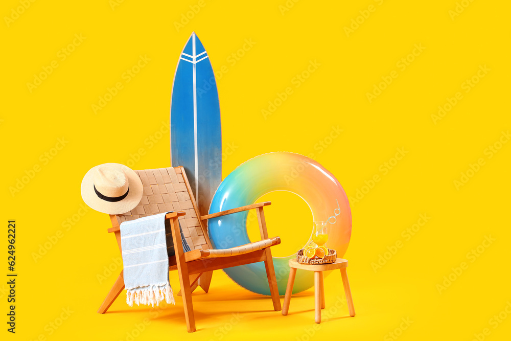 Chair with surfboard, fresh orange juice on stool and beach accessories against yellow background. T