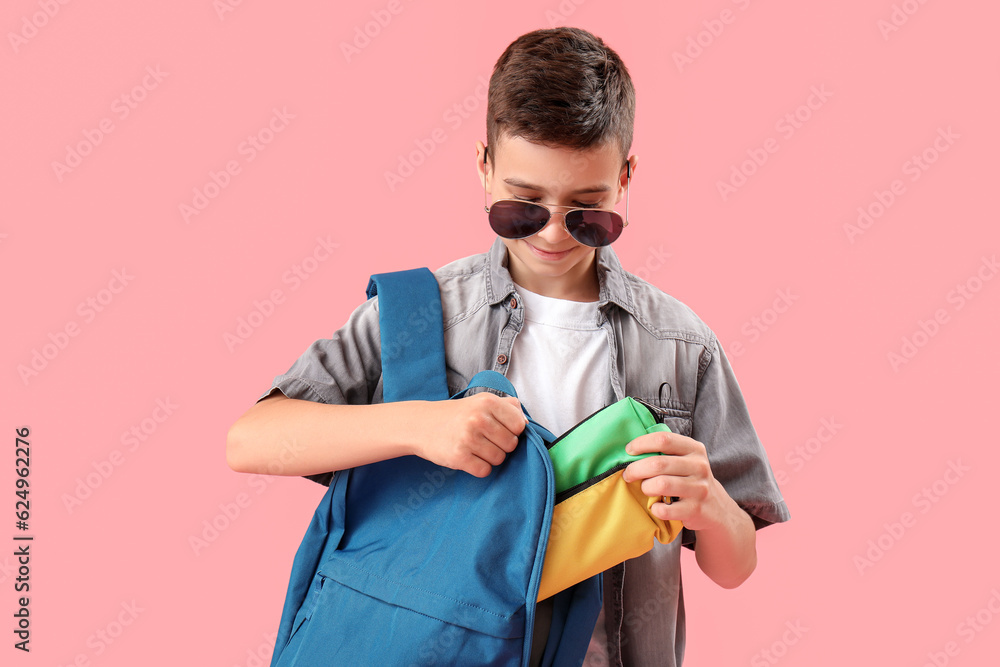 Little schoolboy with backpack and pencil cases on pink background