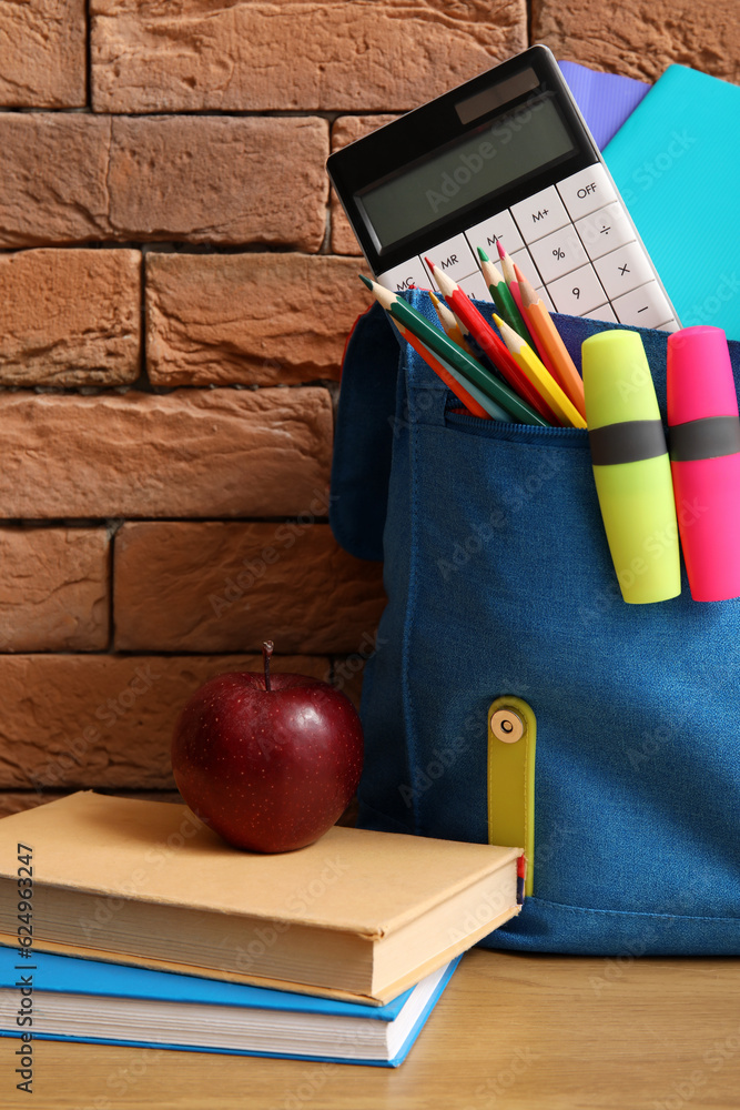 Blue school backpack with different stationery and fresh apple on wooden table near brown brick wall