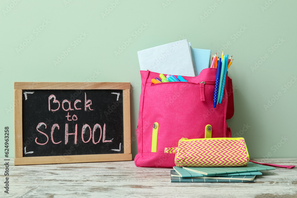 Pink school backpack, stationery and blackboard with text BACK TO SCHOOL on wooden table near pale g