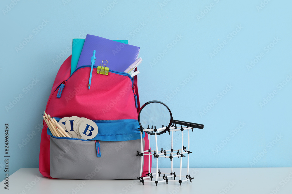 Colorful school backpack with different stationery and molecular model on white table near blue wall