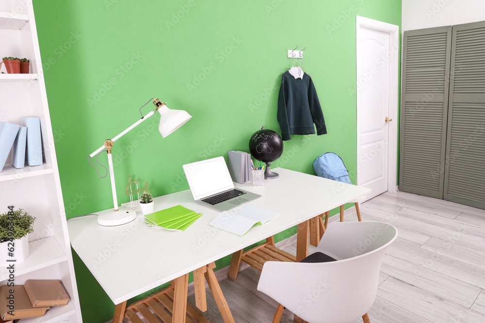 Workplace with desk, laptop computer, lamp and stylish school uniform hanging on green wall