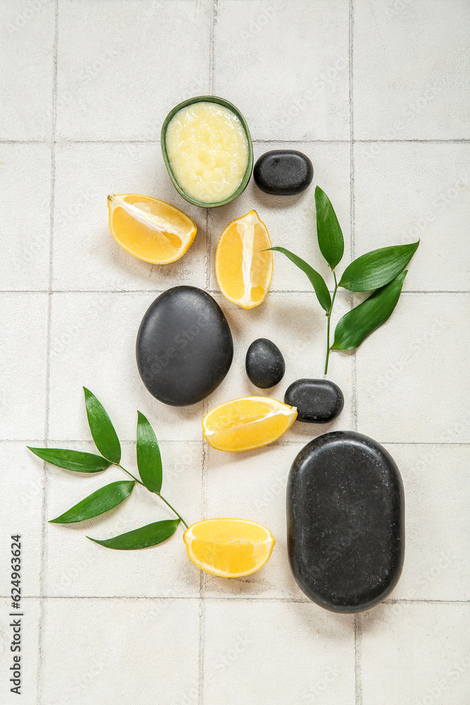 Bowl of lemon body scrub with spa stones and plant branches on white tile background