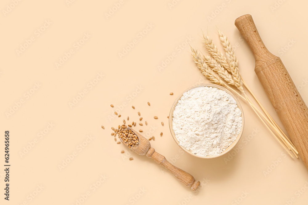 Bowl with wheat flour, spikelets, rolling pin and scoop on beige background