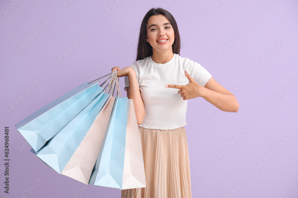 Beautiful young woman pointing at shopping bags on lilac background