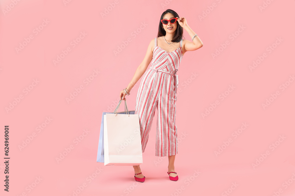 Happy beautiful young Asian woman in sunglasses with shopping bags on pink background