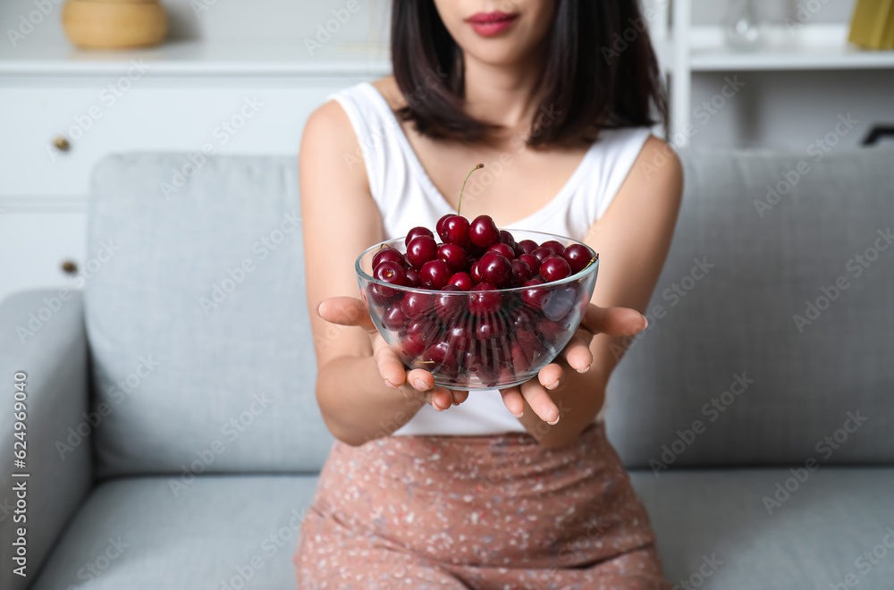 Beautiful young Asian woman with bowl of ripe cherries sitting on sofa in living room