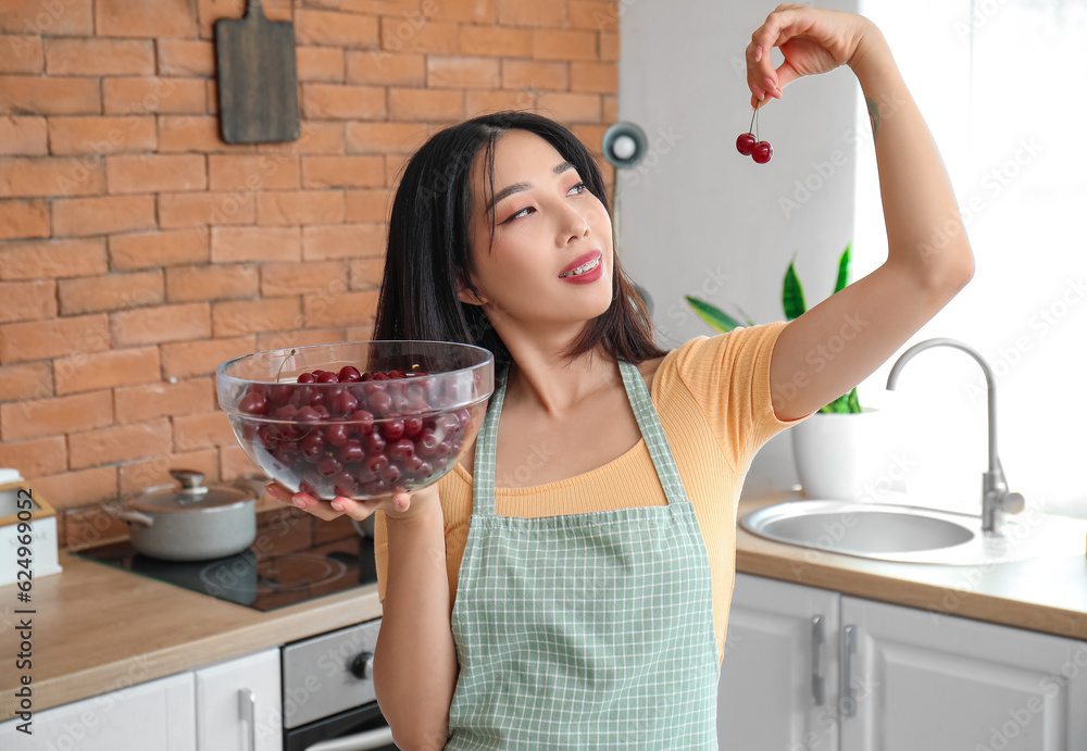 Beautiful young Asian woman in apron with bowl of ripe cherries in kitchen