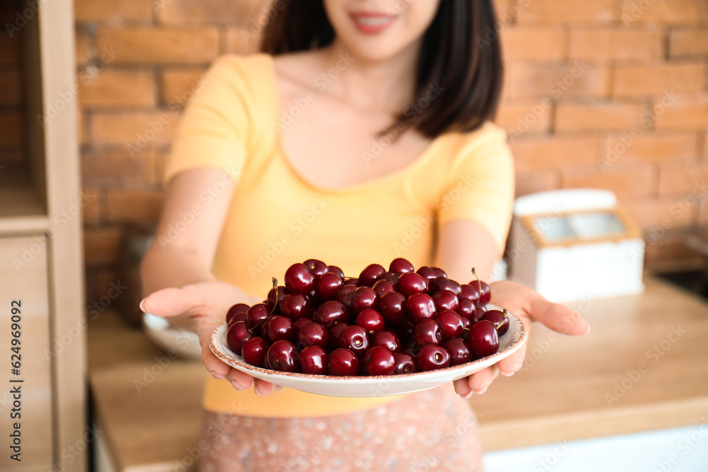 Beautiful young Asian woman with bowl of ripe cherries in kitchen