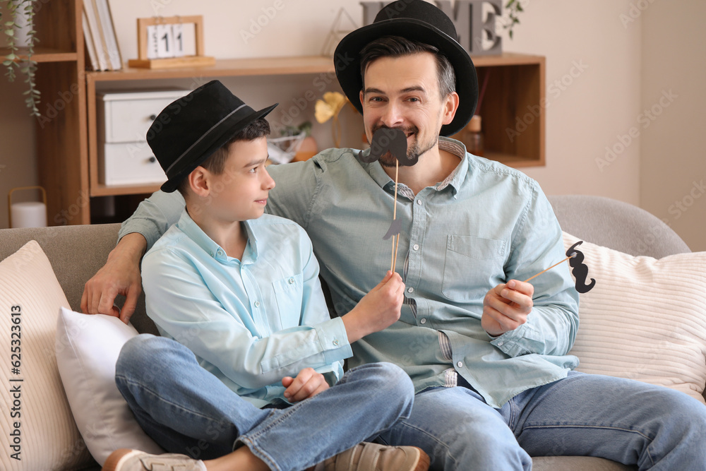 Portrait of father and his little son with paper mustache sitting on sofa