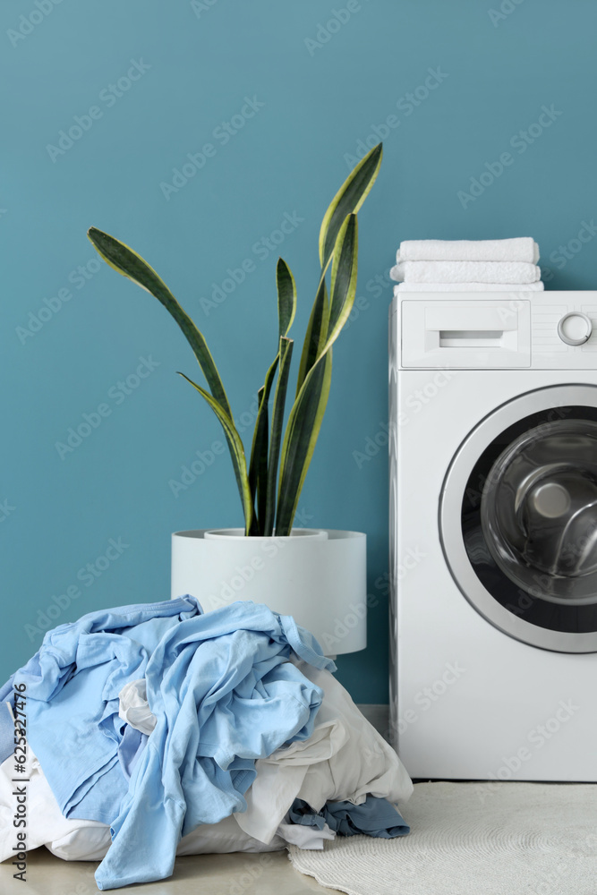 Washing machine, houseplant and pile of dirty laundry near blue wall