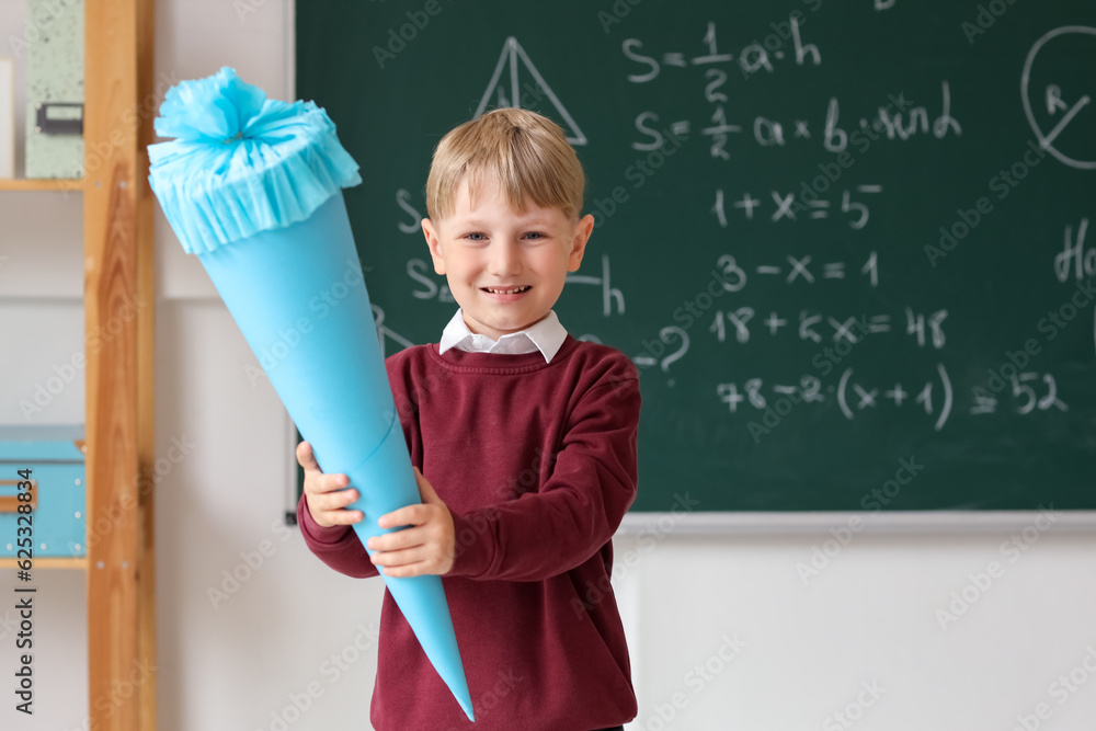 Happy little boy with blue school cone in classroom near blackboard