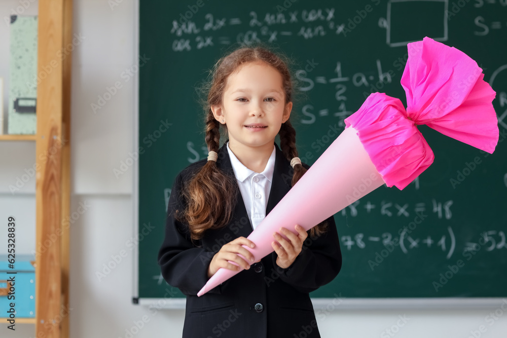 Happy little girl with pink school cone in classroom near blackboard