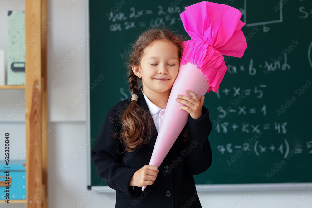 Happy little girl with pink school cone in classroom near blackboard