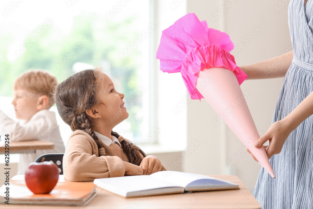 Teacher greeting her happy pupil with pink school cone in classroom