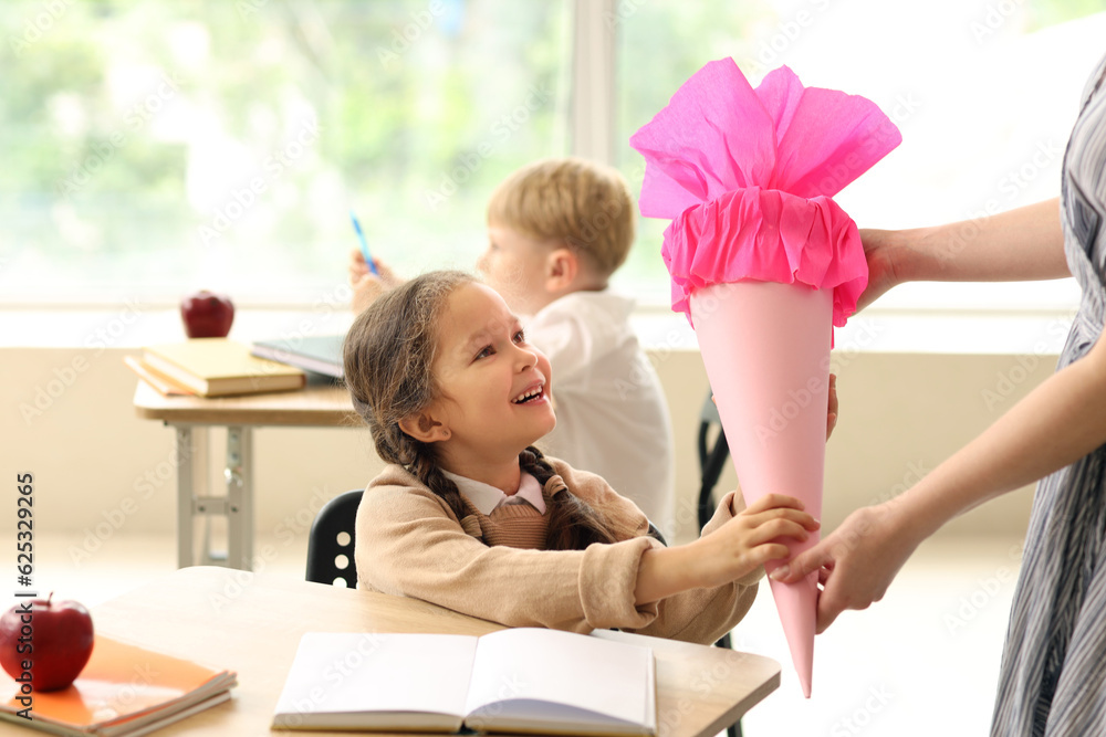 Teacher greeting her happy pupil with pink school cone in classroom