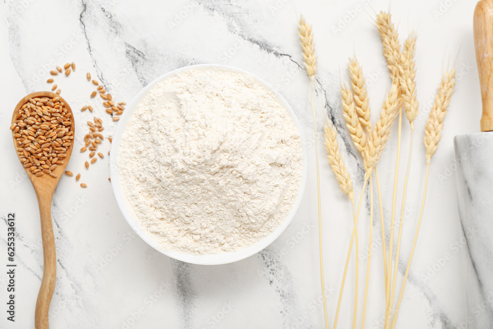 Wheat flour in bowl, spoon with grains and spikelets on white table