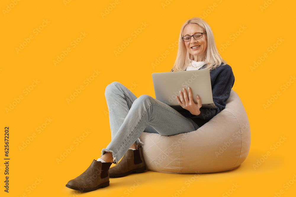 Mature female programmer working with laptop in beanbag on yellow background