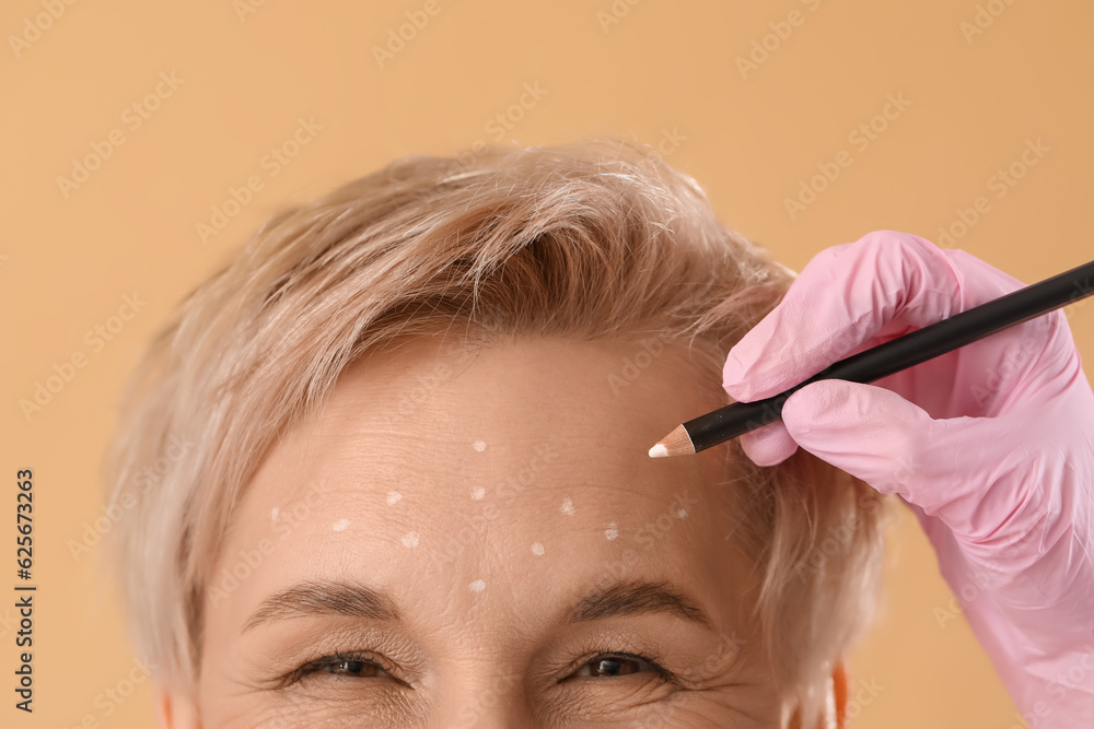 Beautician marking forehead of mature woman for filler injection on beige background, closeup