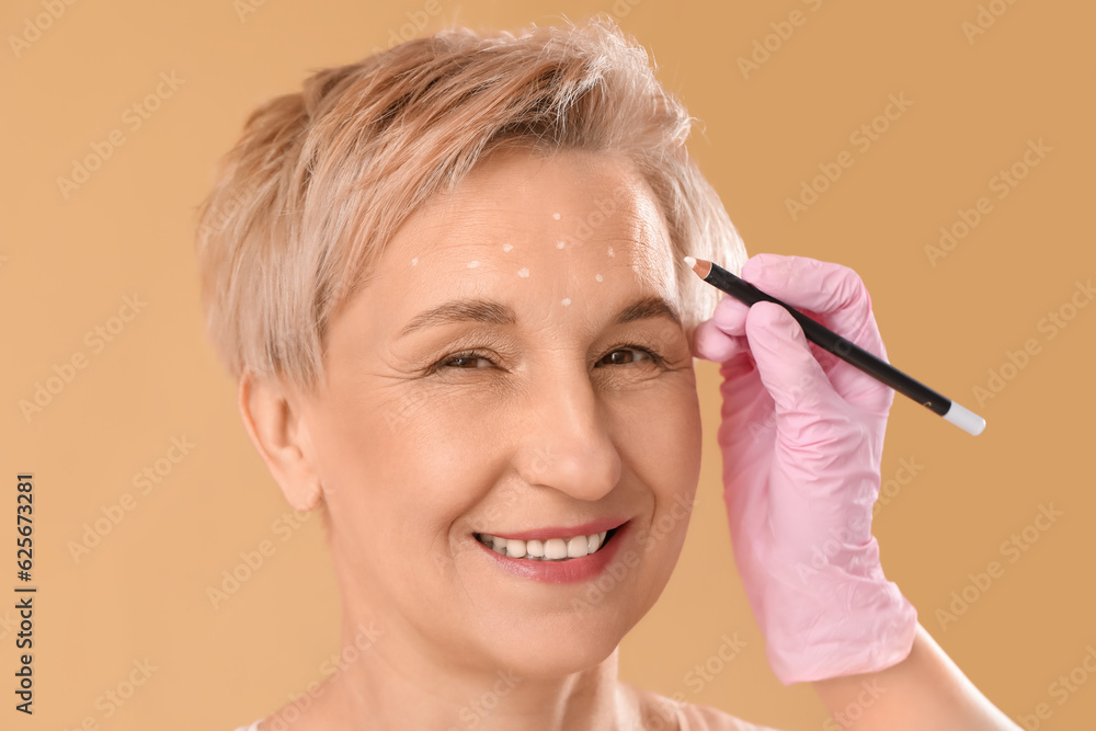 Beautician marking forehead of mature woman for filler injection on beige background, closeup