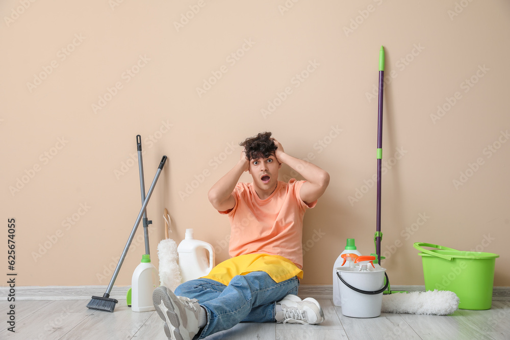 Shocked young man with cleaning supplies sitting near beige wall