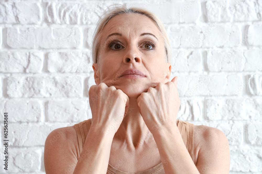 Mature woman doing face building exercise on white brick background, closeup