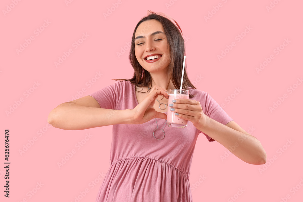 Young pregnant woman with glass of smoothie making heart on pink background