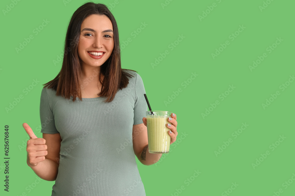 Young pregnant woman with glass of smoothie showing thumb-up on green background