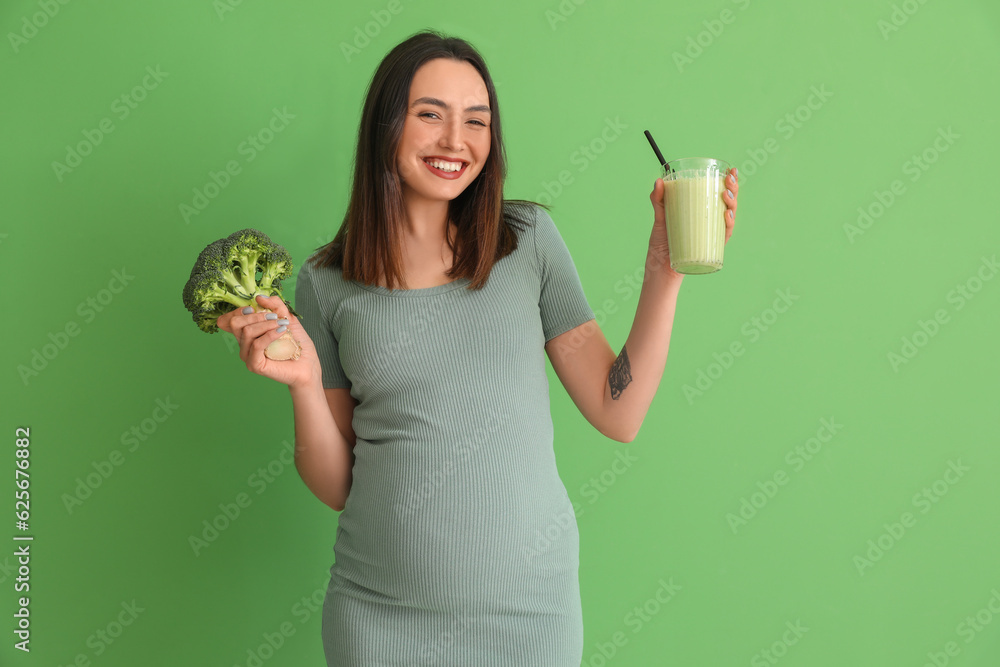 Young pregnant woman with glass of smoothie and broccoli on green background