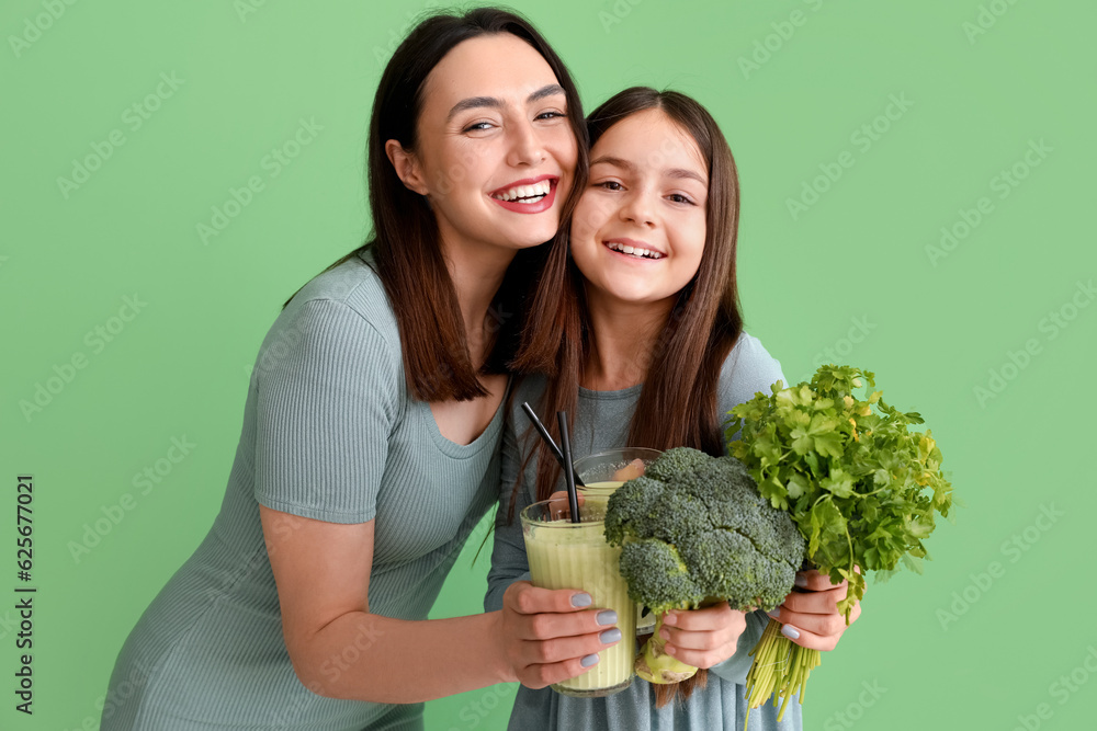 Little girl with her mother and vegetables drinking smoothie on green background